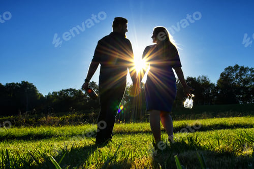 Preview: Male female couple walking through grass with wine bottle glasses at golden hour holding hands