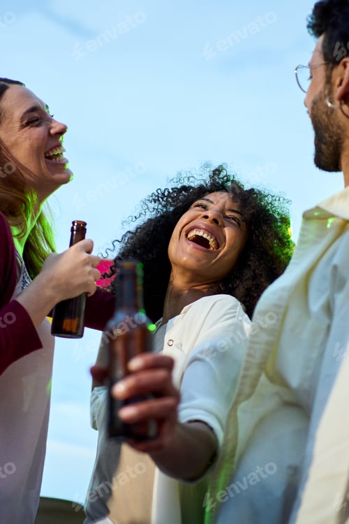 Preview: Focus on excited African woman holding beer bottle with laughing group millennial friends at sunset