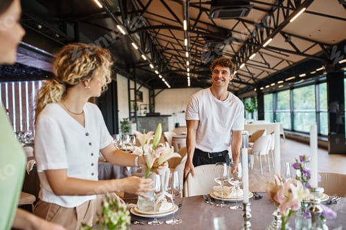 Preview: happy decorator looking at women holding flowers near table with festive setting in event hall