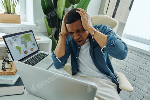 Preview: Frustrated mixed race man holding head in hands while working in the office