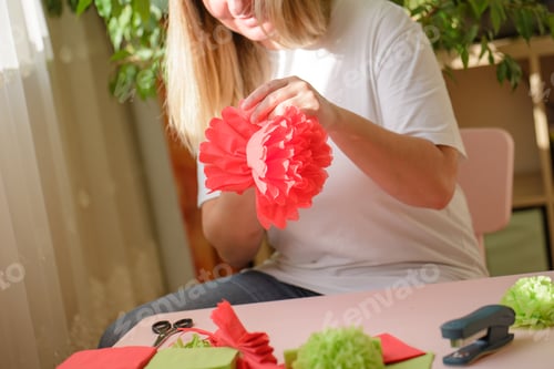 Preview: Women's hands distribute petals on a red blank of napkins for a paper flower in the rays of the sun.