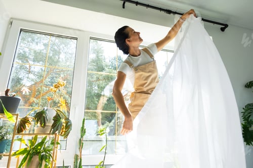 Preview: Woman in apron hangs transparent tulle curtains on large windows in the house inside