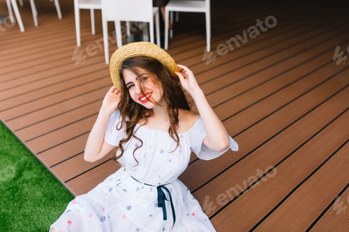 Preview: Pretty girl with long hair in hat is sitting on the wood floor outdoor. She wears a white dress with