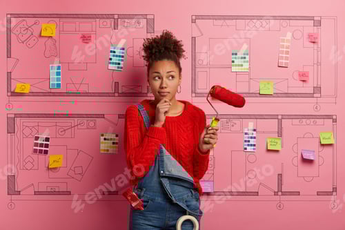 Preview: Home repairing concept. Pensive dark skinned lady holds chin, stands with paint roller, thinks about