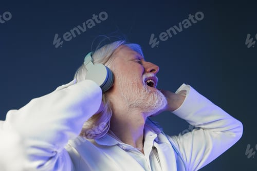 Preview: joyful senior man enjoying music with headphones, singing along, white coat, blue background