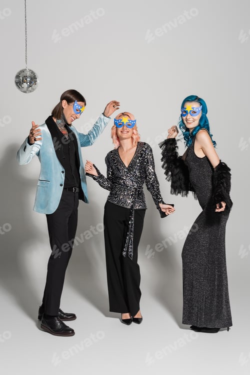 Preview: full length of nonbinary friends in festive clothes and party masks dancing on grey background