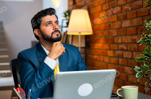Preview: Asian businessman lost in thought while using laptop at desk in cozy workplace with brick wall