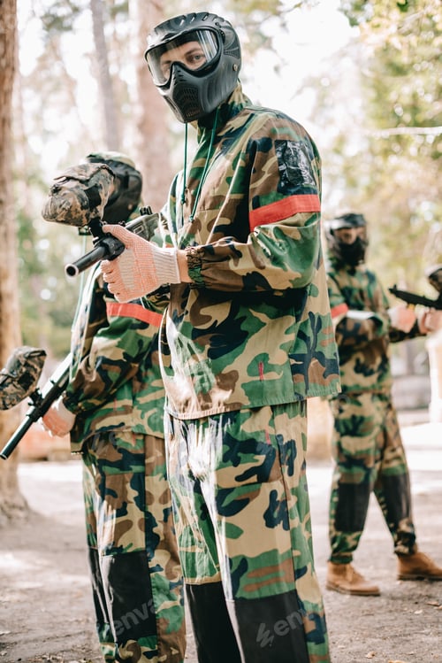 Preview: male paintball player in uniform and goggle mask looking at camera and standing with his team