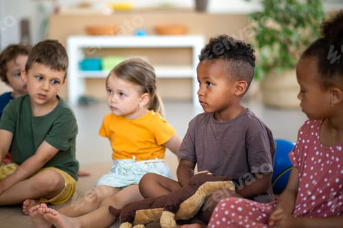Preview: Group of small nursery school children sitting on floor indoors in classroom, listening to teacher