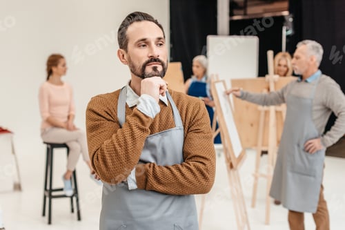 Preview: pensive bearded artist in apron looking away while adult students painting at art class