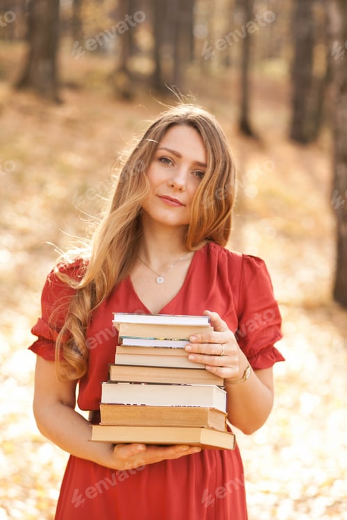 Preview: Beautiful woman in orange dress holding a stack of books in autumn park or forest