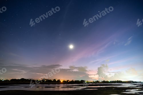 Preview: Blue sky and moon with an orange hue at night over a muddy tidal pool in Lhokseumawe