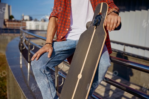 Preview: Closeup man holding skateboard in hand while sitting on steel railing