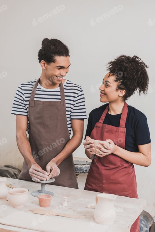 Preview: smiling young african american couple in aprons making clay pots and looking at each other in