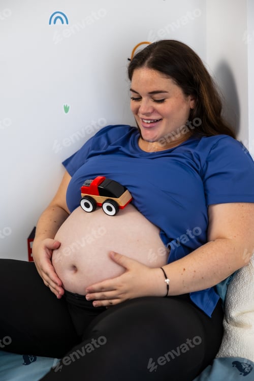 Preview: Pregnant woman smiling with toy car on bare belly in nursery. Lifestyle. Pregnancy.