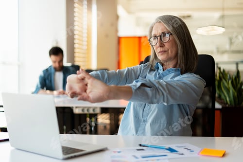 Preview: Mature business woman in casual shirt and glasses, stretching arms after long time sitting at laptop