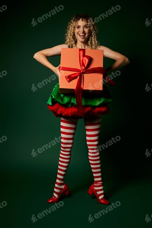 Preview: cheerful blonde woman in new year elf costume holding gift in front of her and smiling at camera
