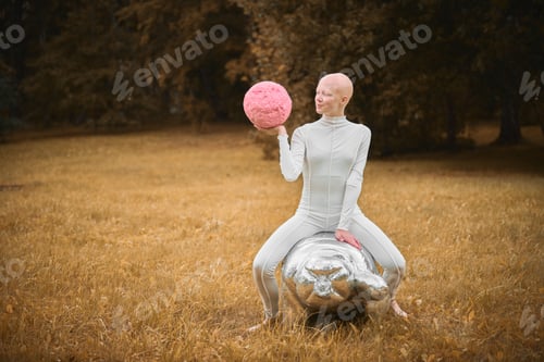 Preview: Young hairless girl with alopecia in white cloth sits on tardigrade figure in fall park