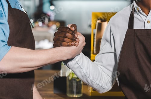 Preview: cropped shot of two owners of coffee shop in aprons shaking hands in coffee shop