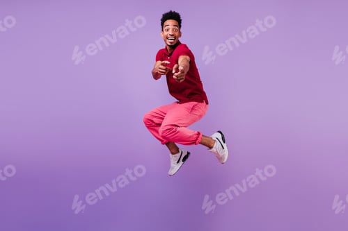 Preview: Carefree young man in white sneakers jumping in studio and pointing finger. Surprised african guy i