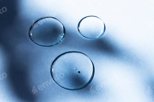 Preview: Transparent drops of a cosmetic product on a blue background with an abstract shadow.