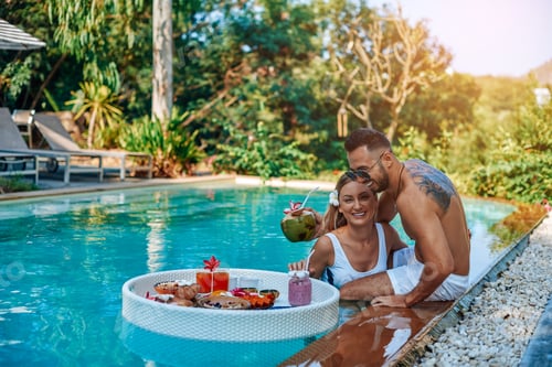 Preview: Laughing couple of travellers swim in a pool with floating table with food
