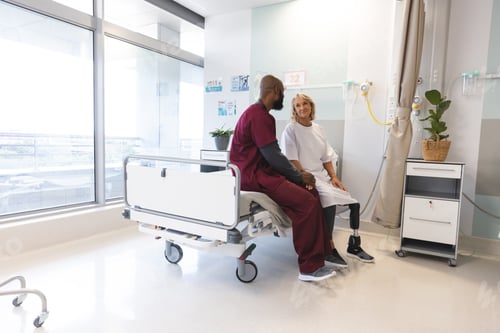 Preview: Senior caucasian female patient with prosthetic leg and african american male doctor sitting on bed