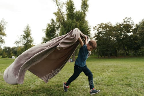 Preview: Cheerful boy running with a flying blanket in the park