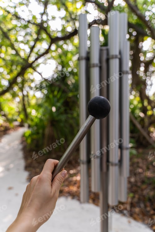 Preview: Female hand with mallet and outdoor chime set in a lush tropical forest