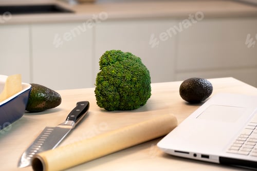 Preview: Kitchen table with broccoli, avocado, knife, baking dish, parchment and computer.