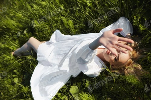 Preview: Girl in a long white dress lies on the grass in a field. Blonde woman in the sun in a light dress