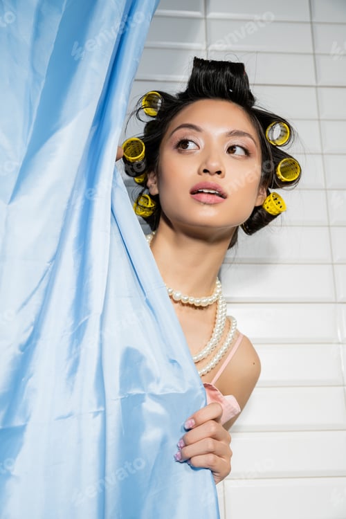 Preview: curious and asian young woman with hair curlers standing in pearl necklace behind blue bathroom