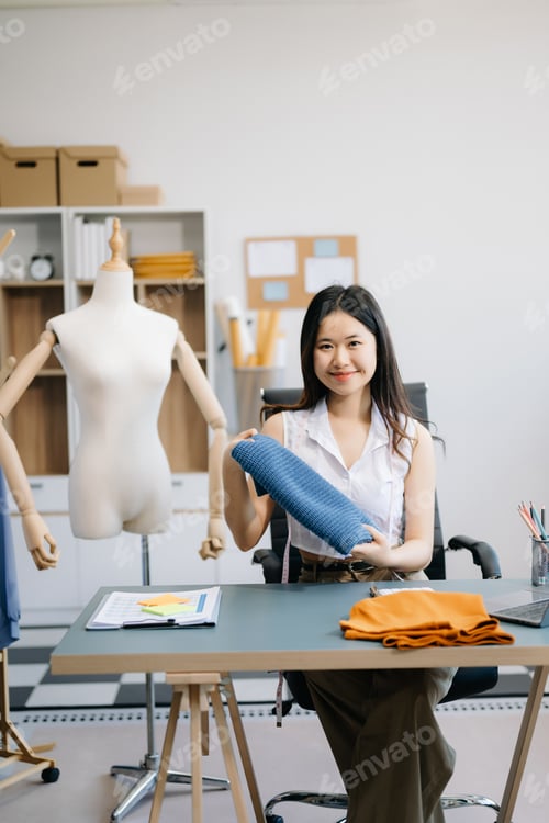 Preview: Asian young woman on desk in office of fashion designer and holds tablet, laptop and smartphone