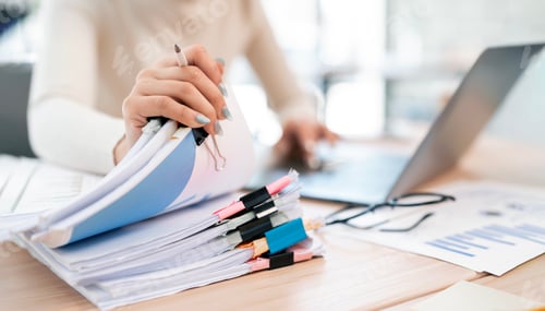 Preview: A woman is seen multitasking, holding a stack of organized documents while working on a laptop.