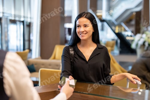 Preview: Happy young businesswoman or female traveler with luggage passing documents to hotel receptionist