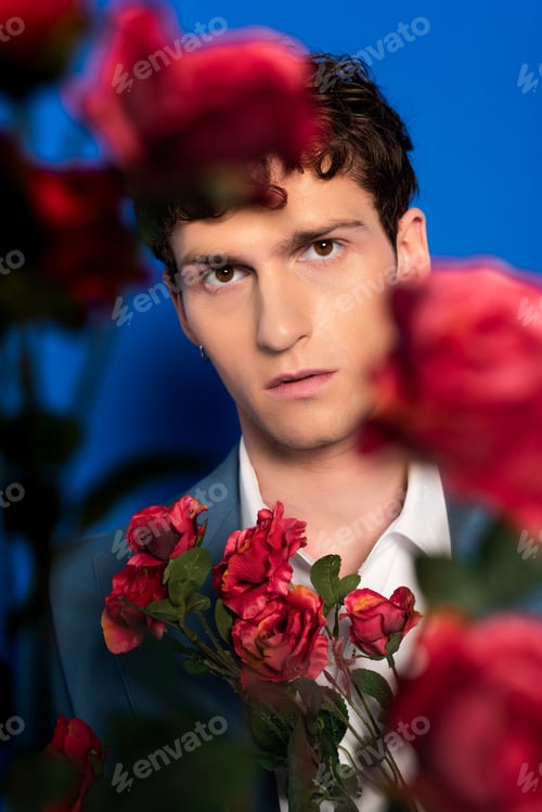 Preview: Trendy young man looking at camera near flowers on blue background