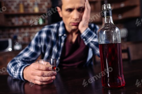 Preview: selective focus of depressed man sitting at table with bottle and glass of alcohol at home