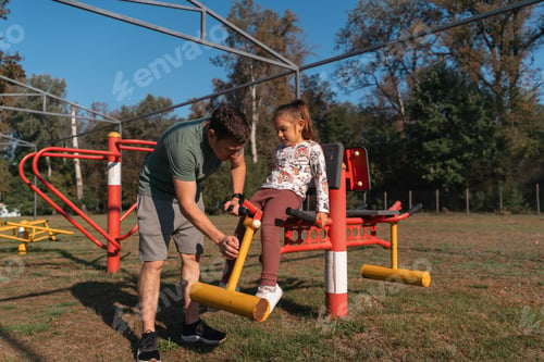Preview: Father and child girl using outdoors workout machine trainer in a park. Healthy active lifestyle