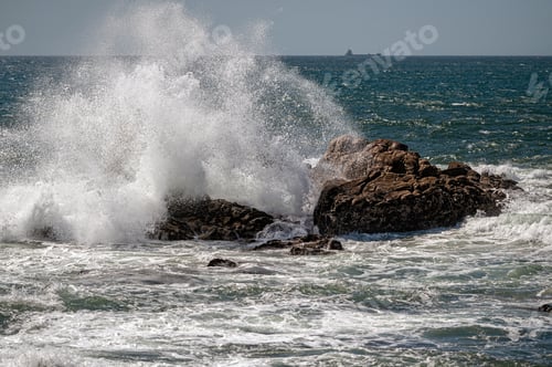 Preview: Powerful ocean waves collide with large rocks along the coast under a clear sky.