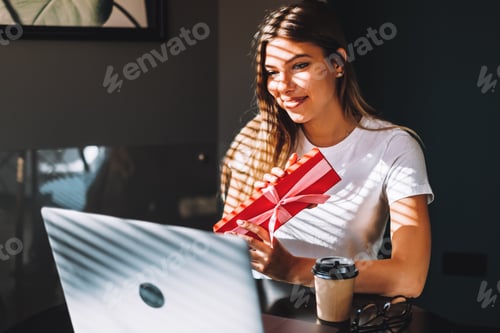 Preview: young woman opening gift in front of laptop during video call or chat, celebrating birthday online