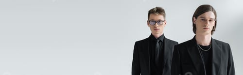 Preview: Young gay looking at camera near boyfriend in formal wear and eyeglasses isolated on grey, banner