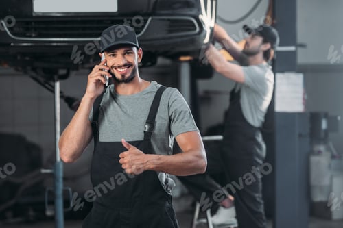 Preview: auto mechanic talking on smartphone and showing thumb up, while colleague working in workshop behind