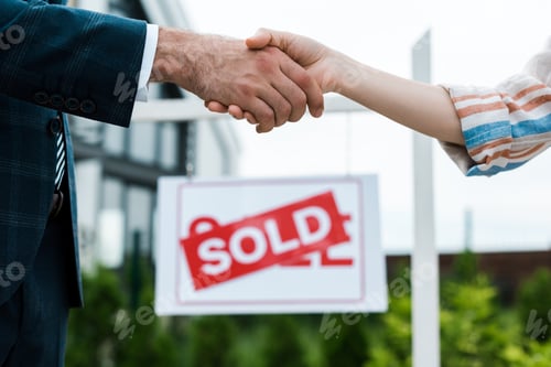 Preview: selective focus of broker and woman shaking hands near board with sold letters