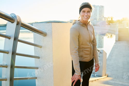 Preview: Young smiling sportswoman in activewear standing on bridge by riverside