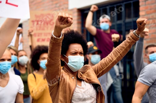 Preview: African American woman wearing face mask while protesting with arms raised on city streets.