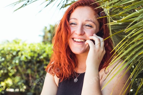 Preview: Close up of smiling attractive girl with redhead talking on mobile phone while standing outdoors