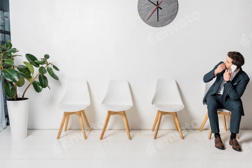 Preview: nervous young businessman talking by smartphone and looking at clock while sitting on chair in line