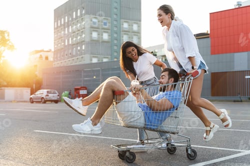 Preview: Three diverse friends having fun and riding shopping cart on parking lot near supermarket
