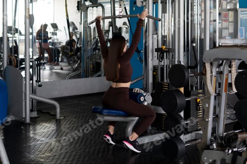 Preview: a young woman lifting weight on a training apparatus in fitness gym