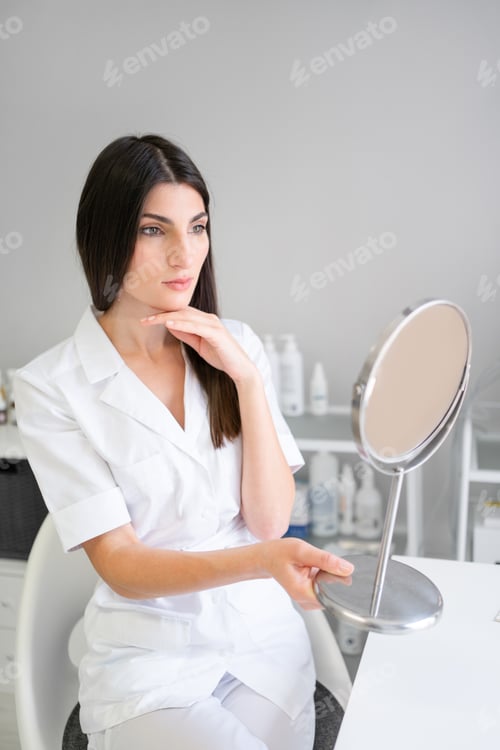 Preview: Confident woman dermatologist holding mirror at desk in clinic